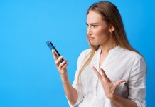 Portrait of young angry woman using her phone, annoyed texting with someone, blue background, close up