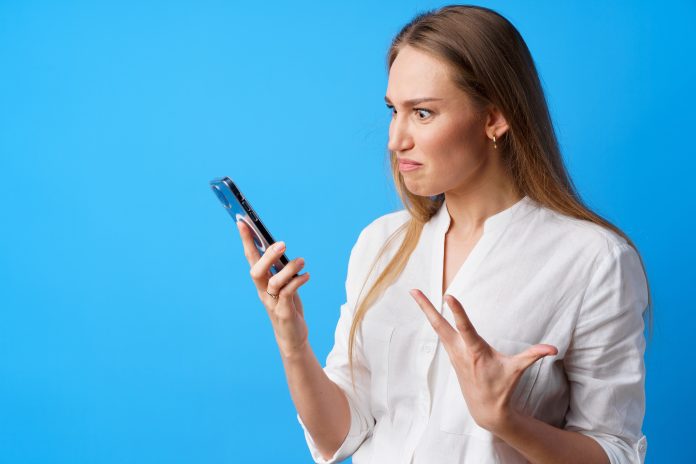 Portrait of young angry woman using her phone, annoyed texting with someone, blue background, close up