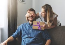 woman giving surprise gift to beloved man at home. closed eyes with hand behind