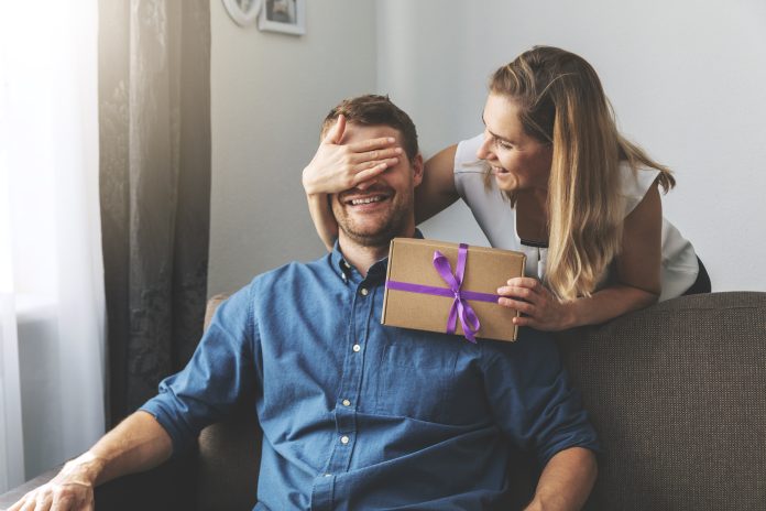woman giving surprise gift to beloved man at home. closed eyes with hand behind